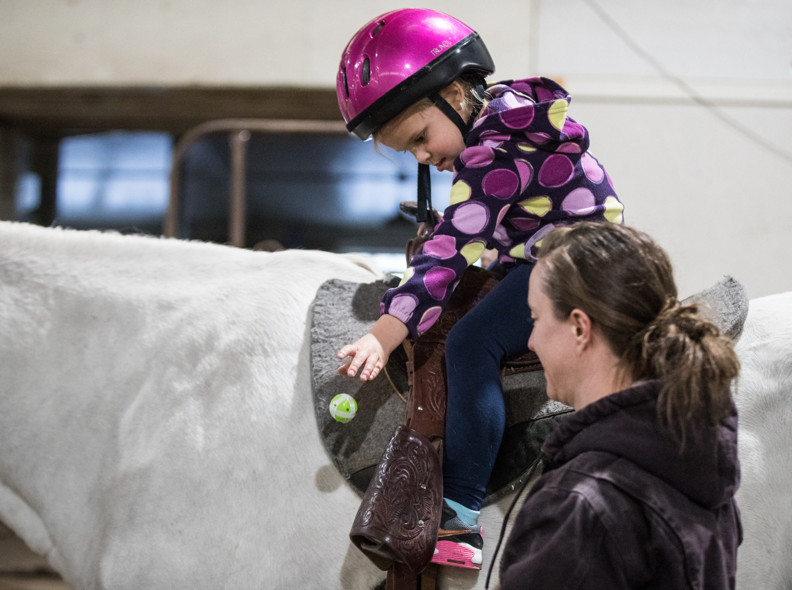 Hazel Guerette leans over the saddle to throw a little green Velcro ball at a target. Her lesson instructor, staff physical therapist Sam Morton, uses this exercise to increase Guerette’s mobility and core strength by moving the target to different spots around Jack’s body, encouraging Hazel to bend and twist.