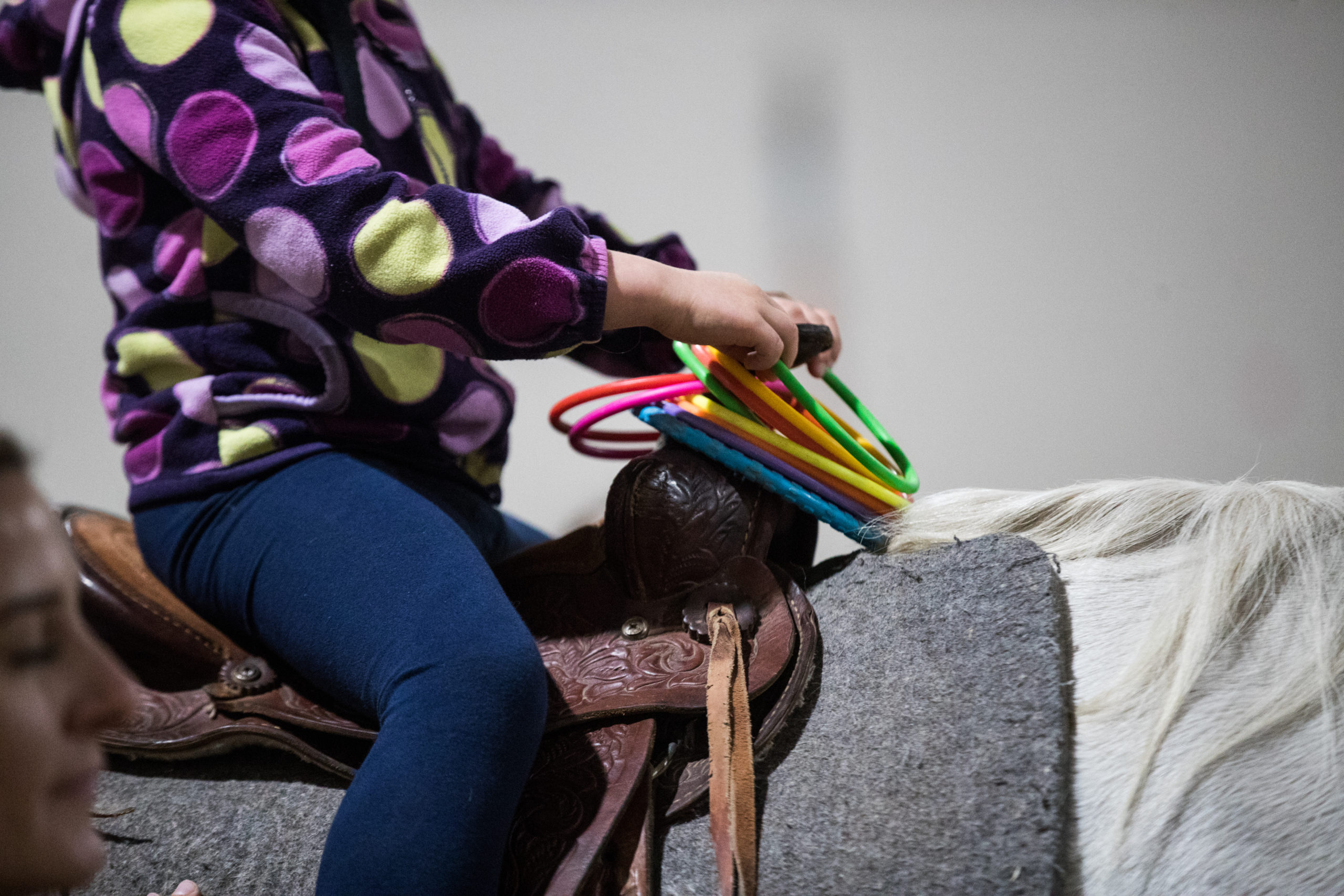 Hazel Guerette puts a green hoop on the saddle horn. This is part of another exercise Sam Morton does with Guerette to improve her core strength and motor skills, similar to the green ball and target. Morton offers her a hoop, encouraging her to lean forward and backward and twist, and then says the color of the hoop. Guerette repeats the color, grabs it and puts it on the saddle horn.